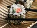 Rolka sushi z łososiem i serkiem, posypana sezamem, trzymana pałeczkami. Czy sushi można jeść na drugi dzień? Tak, jeśli było odpowiednio przechowywane.