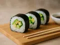 Trzy rolki sushi z awokado do sushi, ułożone na drewnianej desce, obok sosu sojowego i pałeczek.