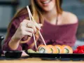 Uśmiechnięta kobieta w ciąży sięga po sushi. Na talerzu rolki z łososiem i awokado, obok wasabi i marynowany imbir.