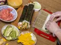 Przygotowanie sushi z warzywami: ryż, awokado, mango, krewetki i marynowany imbir. Ręce w rękawiczkach zwijają rolkę.