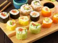 Kolorowe rolki sushi, w tym te z łososiem i awokado, gotowe do zawijania sushi, leżą na drewnianej desce.
