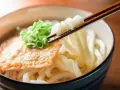 Miseczka z makaronem udon, polana sosem, posypana szczypiorkiem. Pałeczki unoszą porcję grubego makaronu. Pyszne danie, które warto znać pod kątem makaron udon kcal.