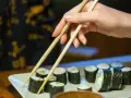 Dłoń podnosi pałeczkami rolkę sushi. Na talerzu ułożone są kawałki sushi, każdy z nich ma około 30 kcal.