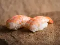 Dwa kawałki sushi z ryżem i różowymi krewetkami do sushi, ułożone na kamiennej powierzchni.