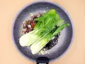 Na patelni smaży się aromatyczny sos z czosnkiem i kawałkami cebuli, a na nim leżą świeże liście sałaty pak choi.