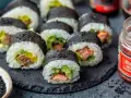 Pyszne sushi z łososiem, awokado i ogórkiem, posypane czarnym sezamem. Obok słoiczek marynowanego imbiru.
