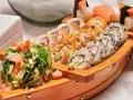 Drewniana łódka pełna sushi, przypominająca o tym, skąd pochodzi sushi. Obok stos białych talerzyków.