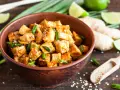 Pokrojone w kostkę, glazurowane tofu z zieloną cebulką i sezamem w misce. Obok limonka, imbir i drewniana łyżka z sezamem.