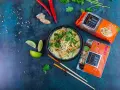 Aksamitna zupa laksa, pełna aromatycznych przypraw i makaronu, to kwintesencja dania tajskie. Obok leżą składniki: papryczki chili, imbir, limonka i opakowania makaronu oraz pasty laksa.