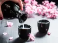 Dłoń nalewa sake z czarnego dzbanka do małej czarki. W tle kwitnące wiśnie.