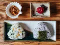 Dwa onigiri z różnym nadzieniem do onigiri: jedno z zielonymi dodatkami i rybkami, drugie z fioletowym ryżem i nori. Obok miseczka orzeszków i marynowana śliwka.