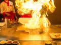 Pokaz teppanyaki z płomieniami buchającymi z płyty grillowej. Kucharz w czerwonym fartuchu dodaje składniki, tworząc widowiskowy spektakl.