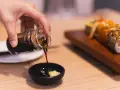 Dłoń nalewa sos sojowy bezglutenowy do miseczki z wasabi. Obok leży sushi.