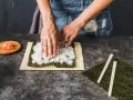 Ręce rozkładają ryż na nori, przygotowując się do zwinięcia sushi. Obok leżą pałeczki i talerzyk z imbirem, idealny dodatek do sushi.