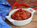 Pikantny sos sambal oelek w białej miseczce, obok czerwonych papryczek chili i niebieskiej tkaniny.
