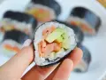 Hosomaki łosoś z awokado, trzymane w dłoni, na talerzu pełnym sushi.