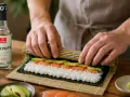 Ręce zwijają sushi z łososiem i awokado. Obok butelka octu ryżowego do sushi.