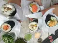 Ręce trzymają cztery pyszne temaki zawinięte w papier. Widać różne nadzienia: łososia, awokado, warzywa i rybę.