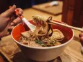 Ręka trzyma pałeczkami porcję parującego ramenu z makaronem i grzybami. Oto jak jesc ramen!