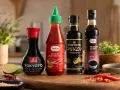 Na drewnianej desce stoją cztery butelki sosów: Vifon Tokyoto Soy Sauce, TaoTao Sriracha Sos Chili Ostry, Tokyoto Ponzu (cytrusowy sos sojowy) i TaoTao Sos Teriyaki Słodki. Obok leżą limonki i papryczki chili.