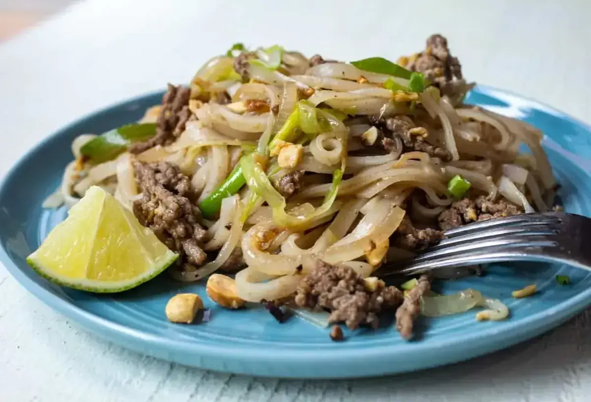 Pyszny pad thai z wołowiną, makaronem ryżowym, orzeszkami ziemnymi i ćwiartką limonki na niebieskim talerzu.