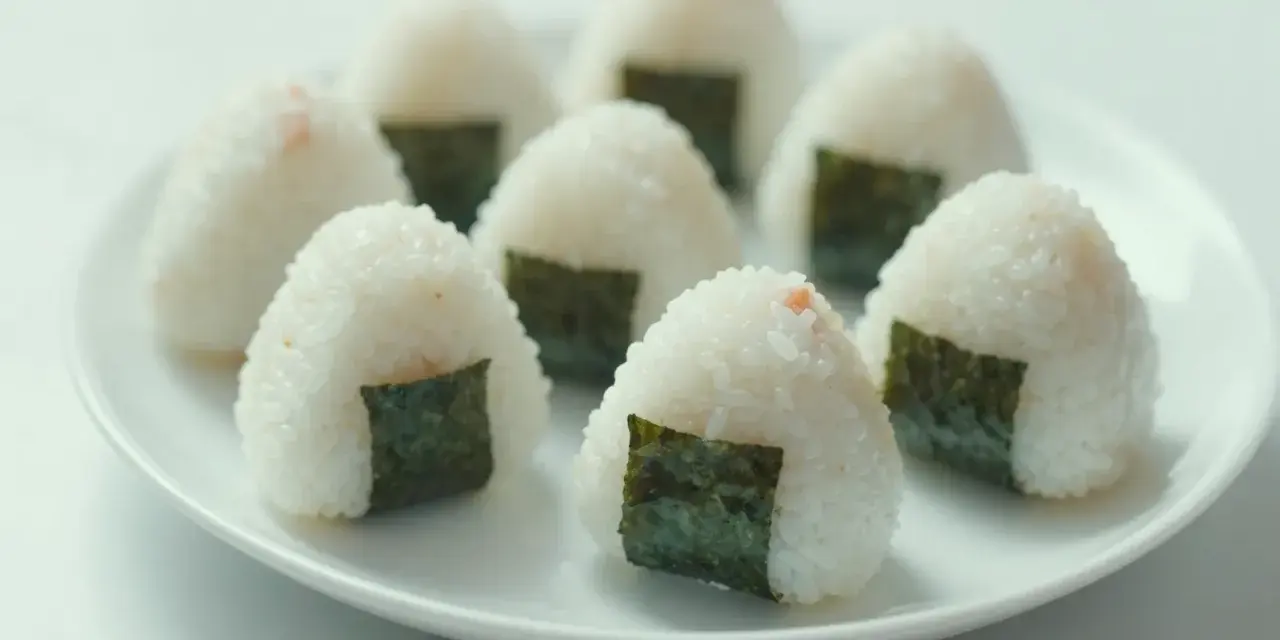 Kilka pysznych onigiri z łososiem, udekorowanych kawałkami nori, ułożonych na białym talerzu.