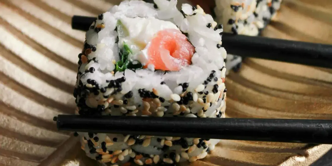 Rolka sushi z łososiem i serkiem, posypana sezamem, trzymana pałeczkami. Czy sushi można jeść na drugi dzień? Tak, jeśli było odpowiednio przechowywane.