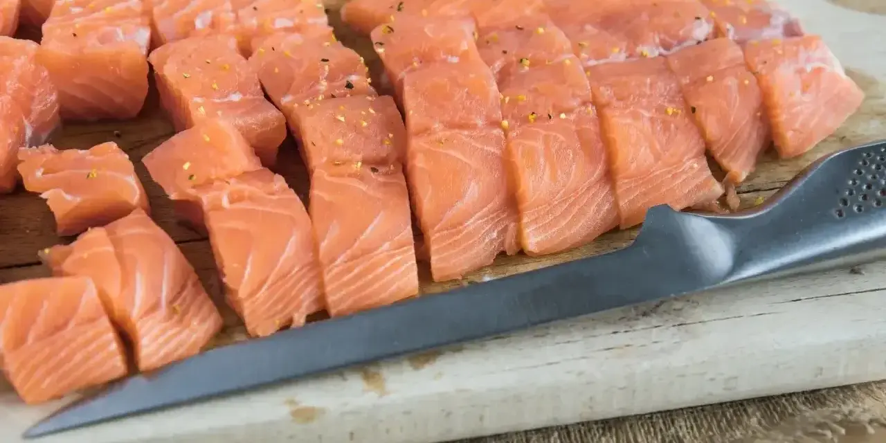 Pokrojona w kostkę i plastry świeża ryba do sushi, przyprawiona, leży na desce obok noża.