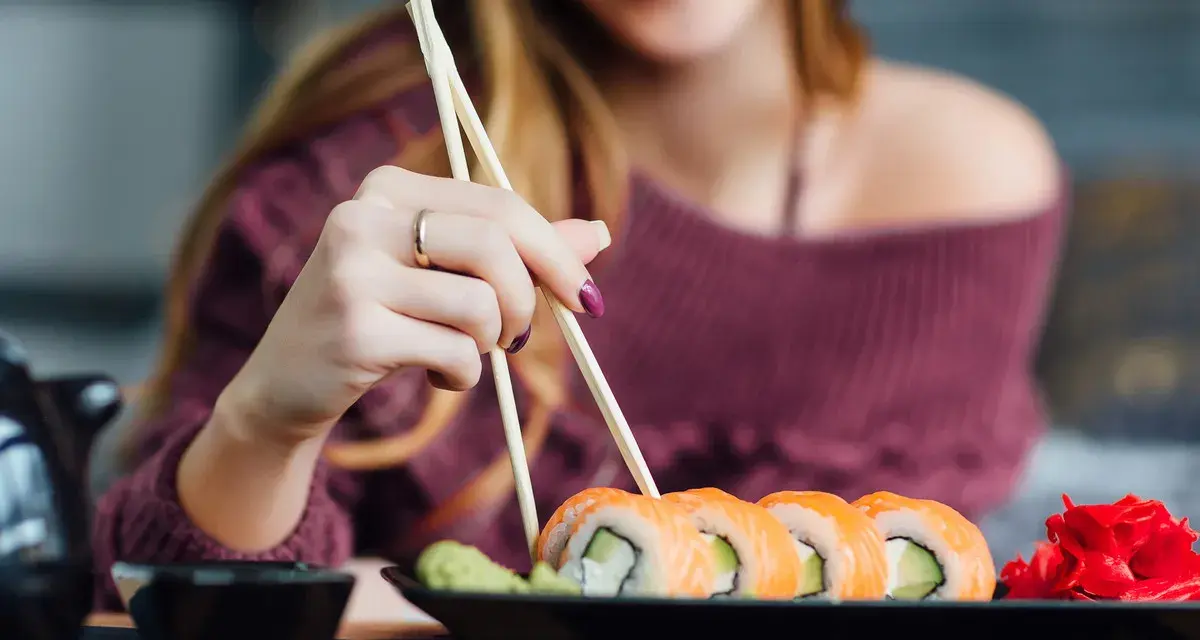 Uśmiechnięta kobieta w ciąży sięga po sushi. Na talerzu rolki z łososiem i awokado, obok wasabi i marynowany imbir.