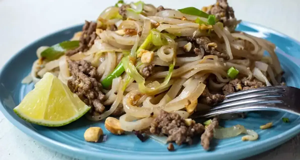Pyszny pad thai z wołowiną, makaronem ryżowym, orzeszkami ziemnymi i ćwiartką limonki na niebieskim talerzu.