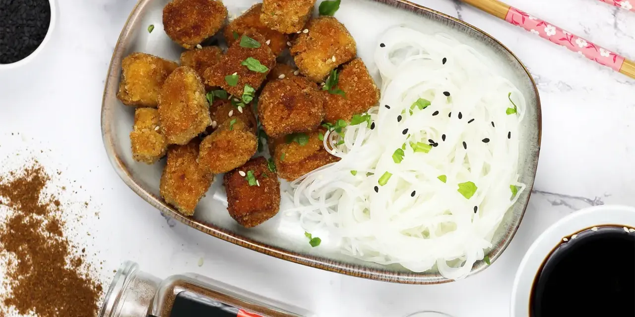 Chrupiące tofu w tempurze z makaronem ryżowym, posypane sezamem i szczypiorkiem. Obok sos sojowy i przyprawy.