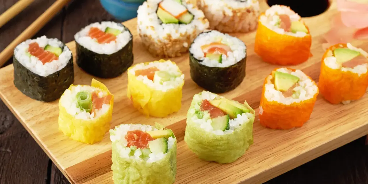 Kolorowe rolki sushi, w tym te z łososiem i awokado, gotowe do zawijania sushi, leżą na drewnianej desce.