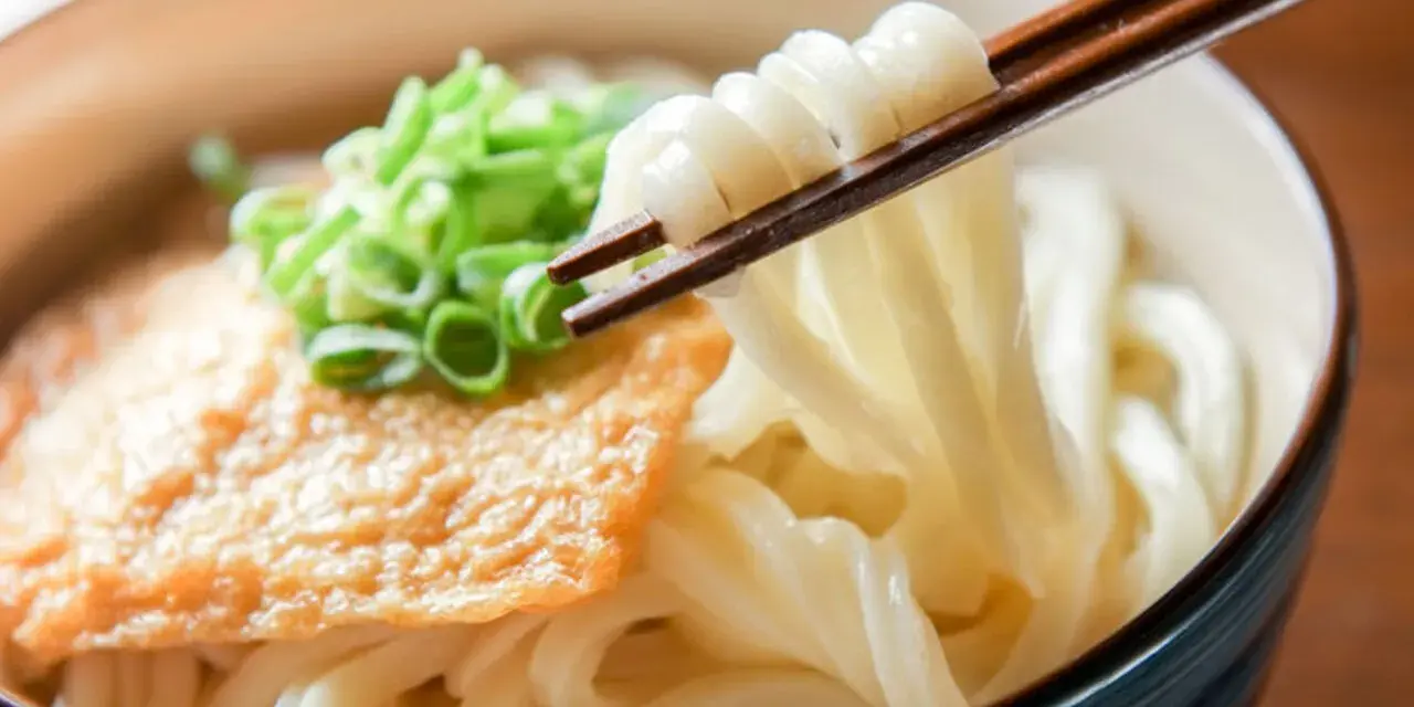 Miseczka z makaronem udon, polana sosem, posypana szczypiorkiem. Pałeczki unoszą porcję grubego makaronu. Pyszne danie, które warto znać pod kątem makaron udon kcal.