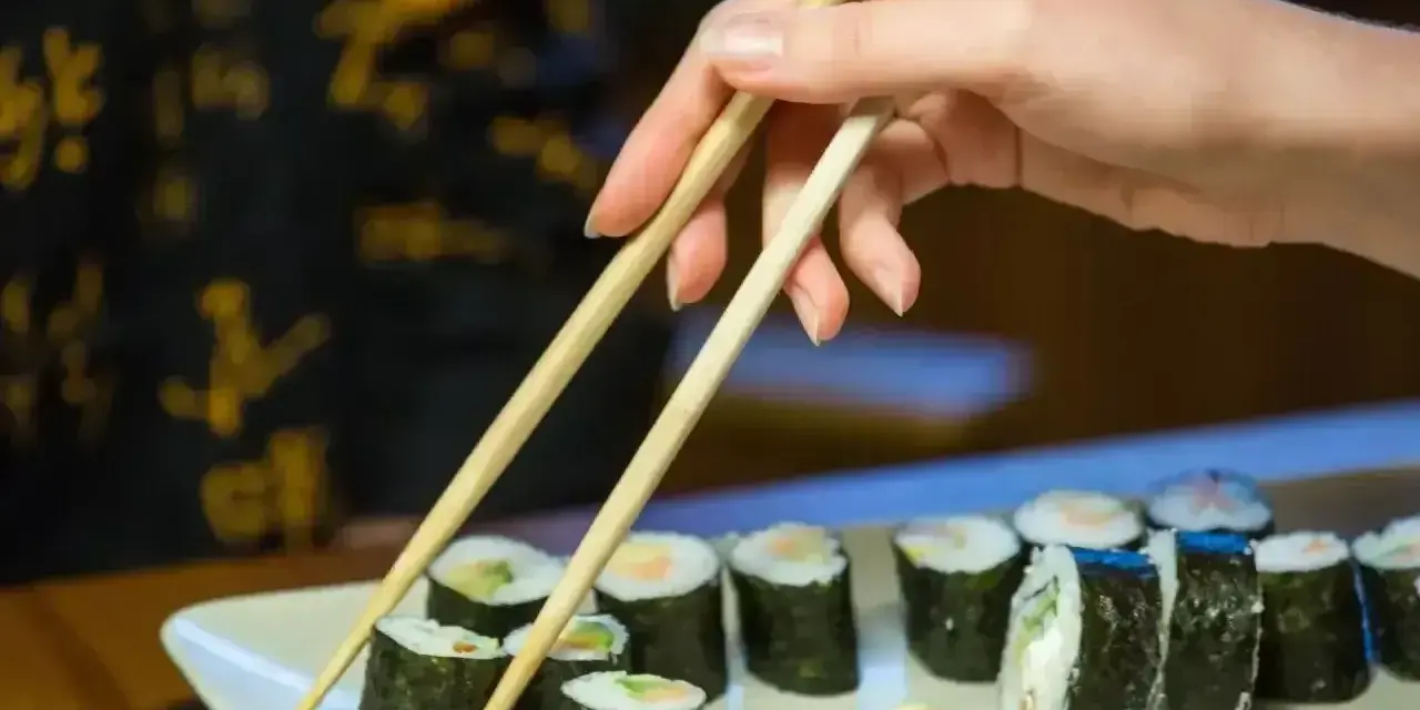 Dłoń podnosi pałeczkami rolkę sushi. Na talerzu ułożone są kawałki sushi, każdy z nich ma około 30 kcal.