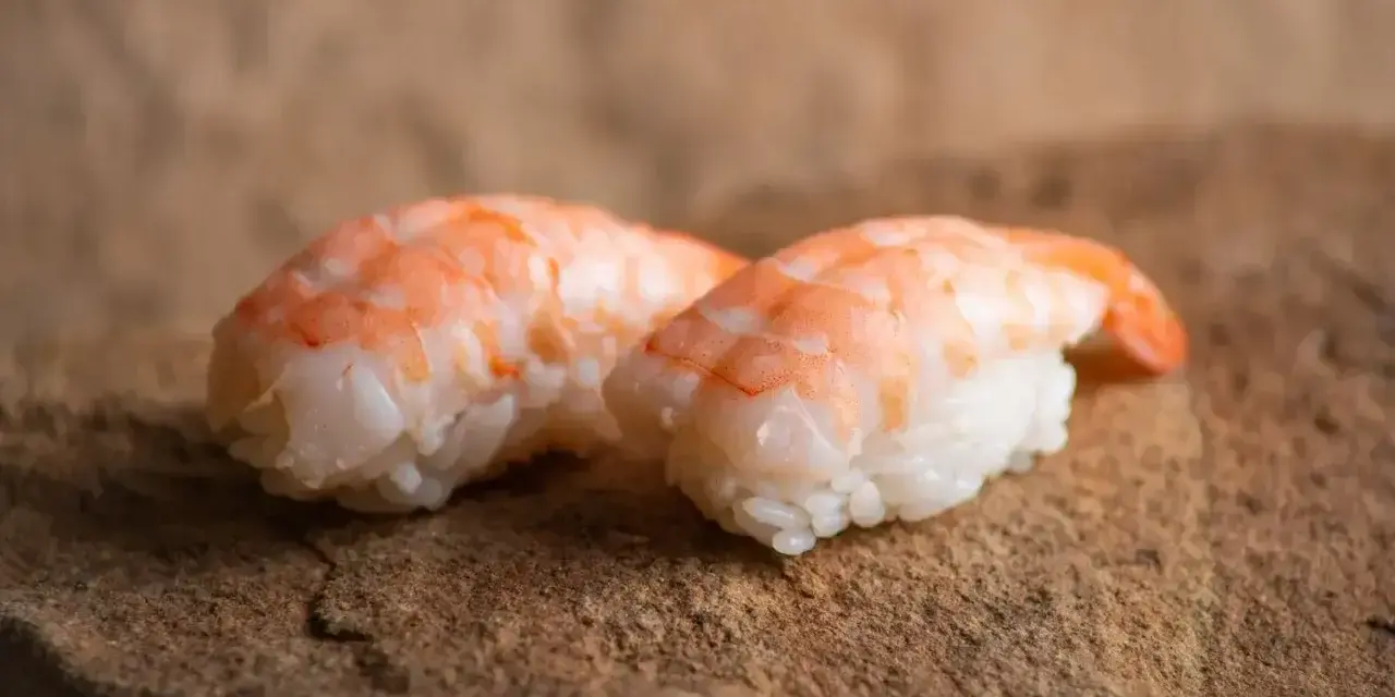 Dwa kawałki sushi z ryżem i różowymi krewetkami do sushi, ułożone na kamiennej powierzchni.
