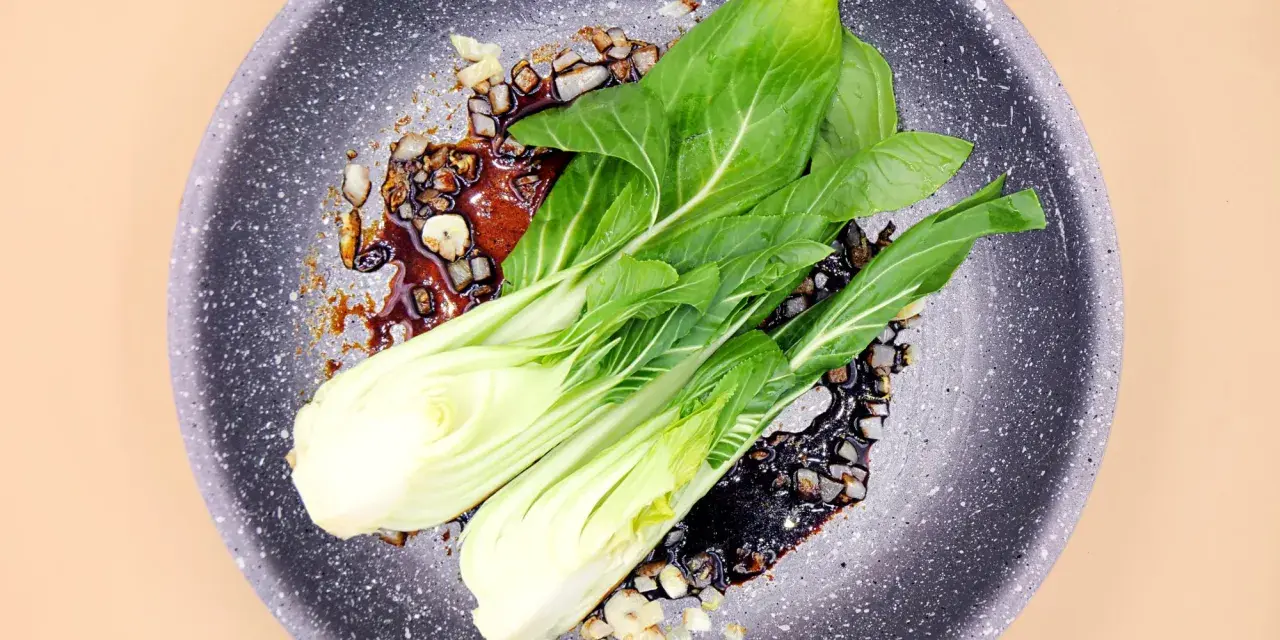 Na patelni smaży się aromatyczny sos z czosnkiem i kawałkami cebuli, a na nim leżą świeże liście sałaty pak choi.