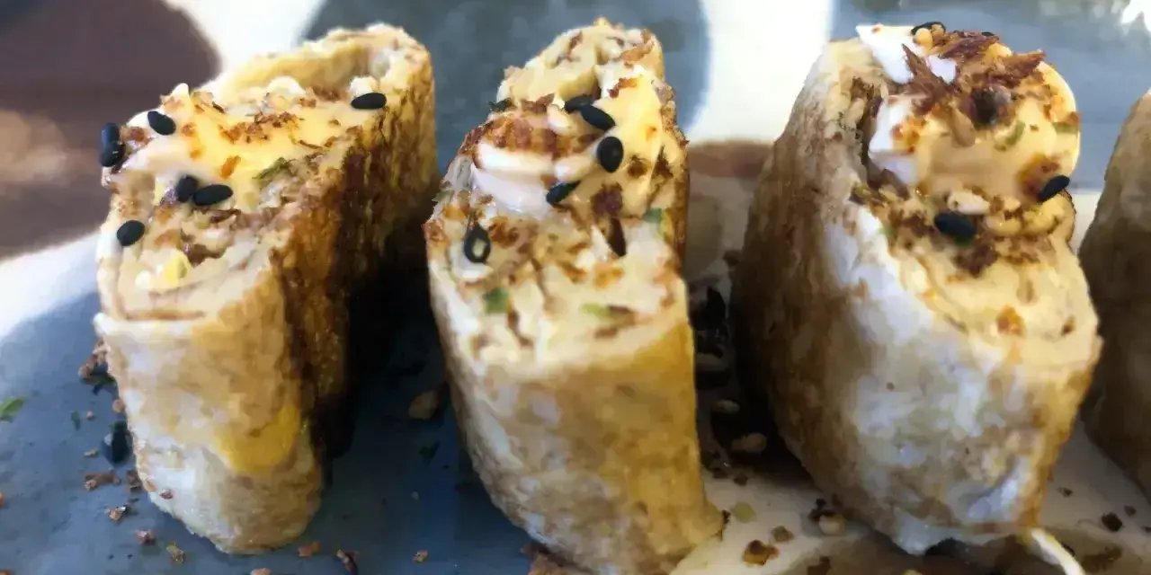 Trzy kawałki sushi z jajkiem, posypane czarnym sezamem i chrupiącymi dodatkami.