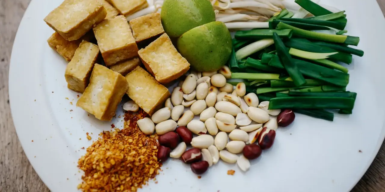 Na talerzu składniki do pad thai: smażone tofu, orzeszki ziemne, szczypiorek, kiełki fasoli mung, limonki i płatki chili. Gotowy sos pad thai czeka!