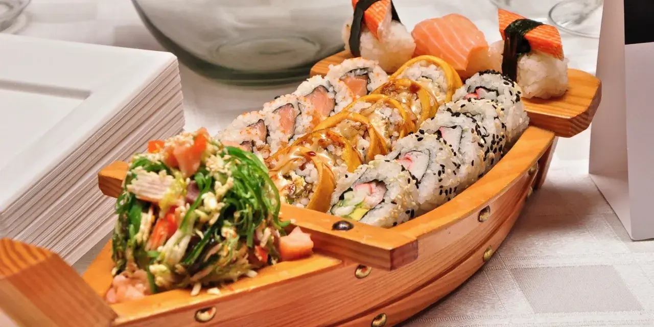 Drewniana łódka pełna sushi, przypominająca o tym, skąd pochodzi sushi. Obok stos białych talerzyków.