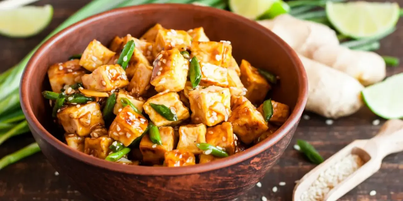 Pokrojone w kostkę, glazurowane tofu z zieloną cebulką i sezamem w misce. Obok limonka, imbir i drewniana łyżka z sezamem.