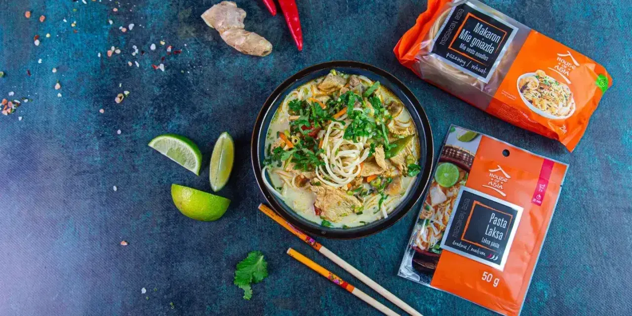 Aksamitna zupa laksa, pełna aromatycznych przypraw i makaronu, to kwintesencja dania tajskie. Obok leżą składniki: papryczki chili, imbir, limonka i opakowania makaronu oraz pasty laksa.