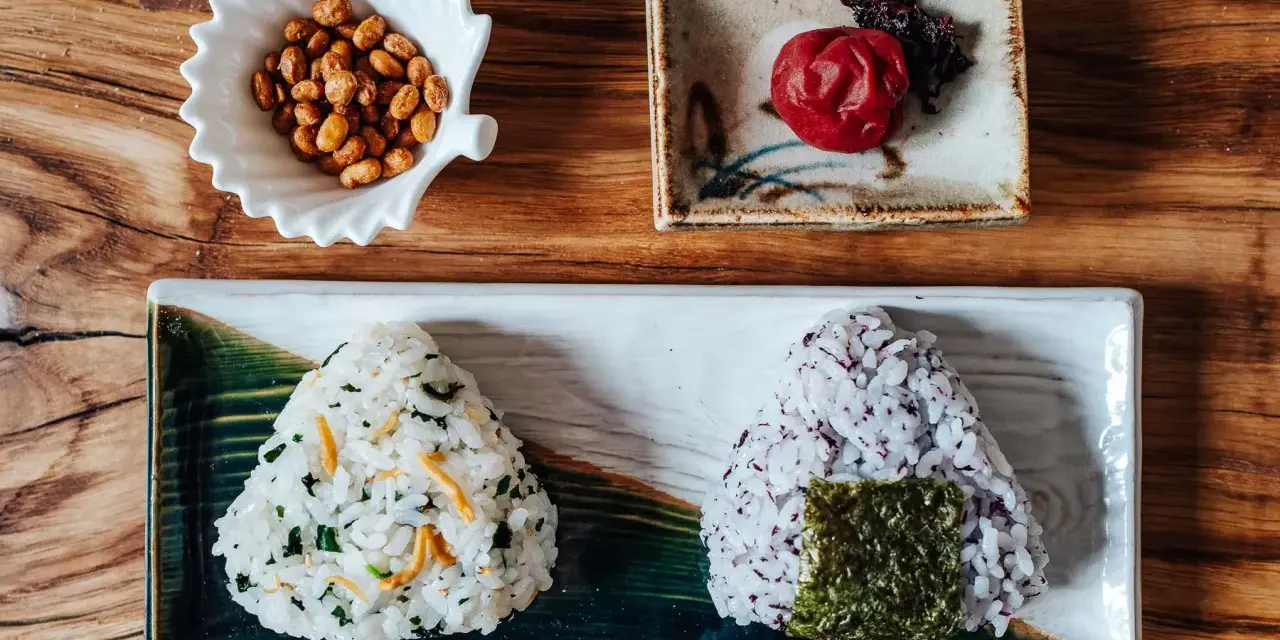 Dwa onigiri z różnym nadzieniem do onigiri: jedno z zielonymi dodatkami i rybkami, drugie z fioletowym ryżem i nori. Obok miseczka orzeszków i marynowana śliwka.