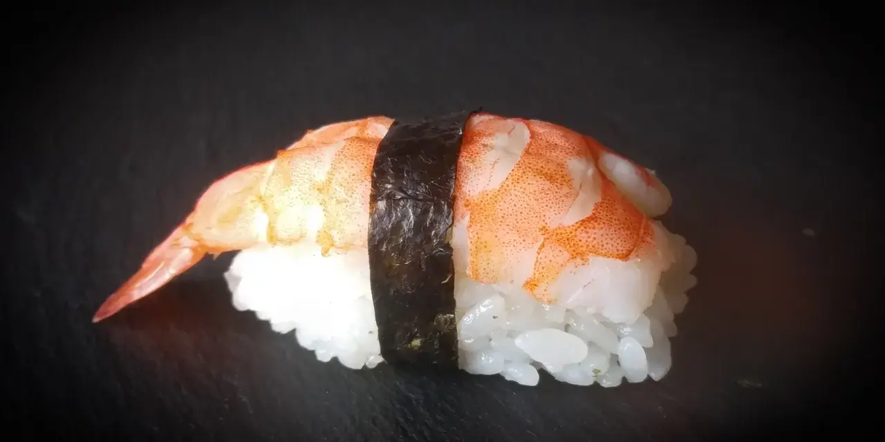 Nigiri ebi, czyli sushi z krewetką, na czarnym tle. Krewetka jest ułożona na ryżu, a całość spięta paskiem nori.