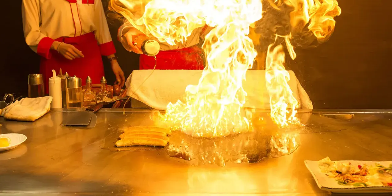 Pokaz teppanyaki z płomieniami buchającymi z płyty grillowej. Kucharz w czerwonym fartuchu dodaje składniki, tworząc widowiskowy spektakl.