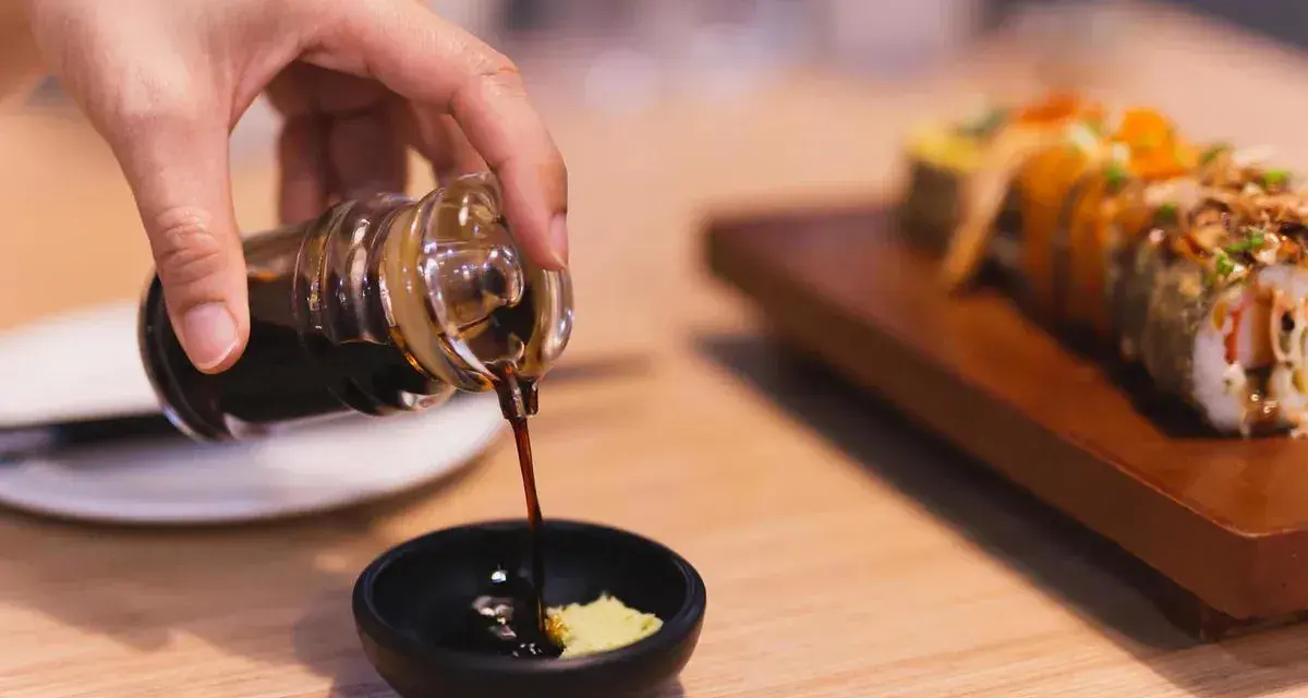 Dłoń nalewa sos sojowy bezglutenowy do miseczki z wasabi. Obok leży sushi.