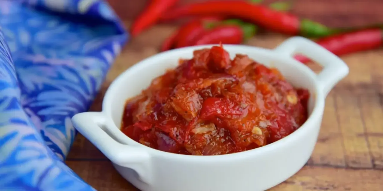Pikantny sos sambal oelek w białej miseczce, obok czerwonych papryczek chili i niebieskiej tkaniny.