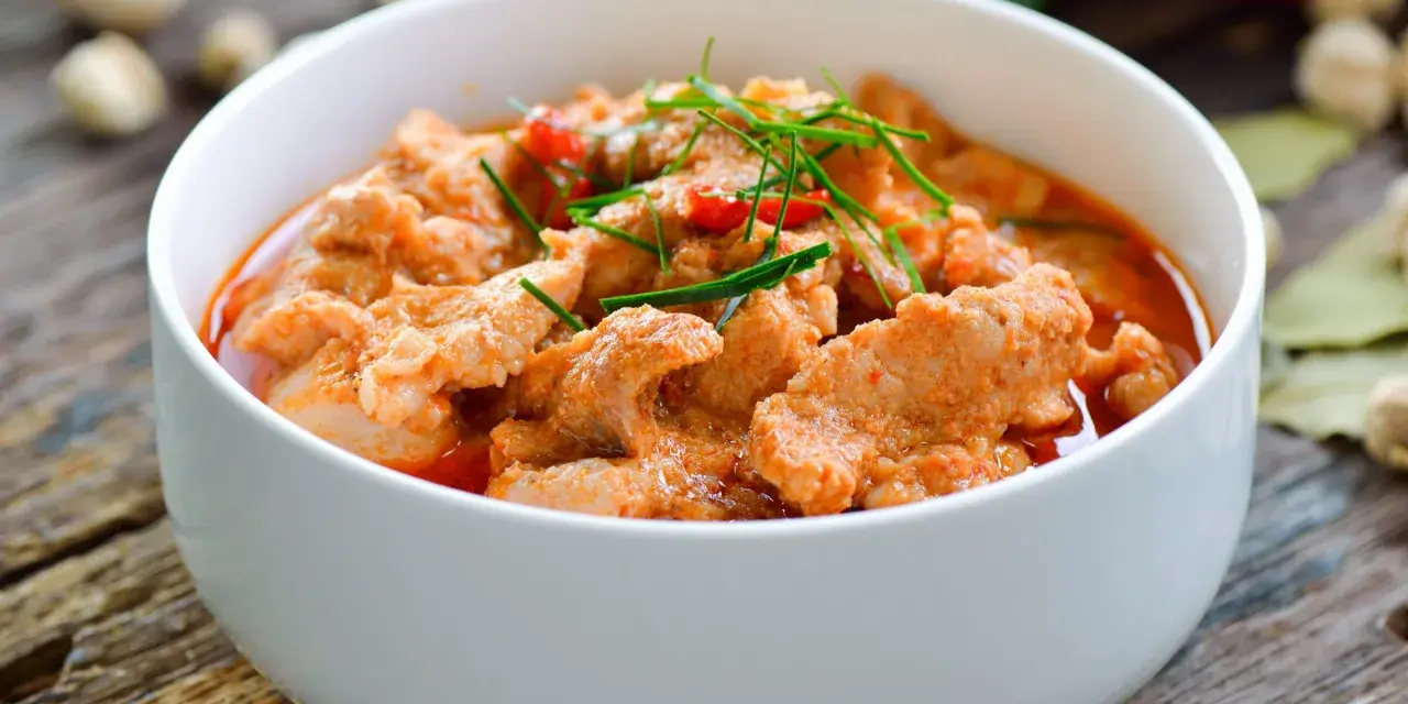 Aromaticzne curry z wieprzowiną w białej misce, ozdobione zieloną szczypiorkiem i czerwonymi papryczkami chili.