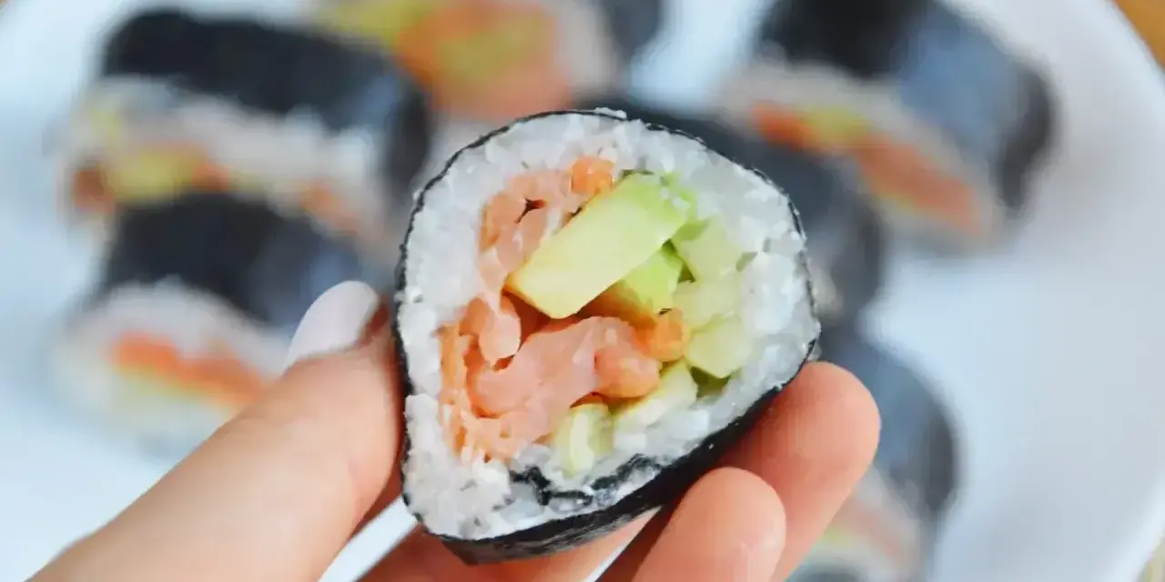 Hosomaki łosoś z awokado, trzymane w dłoni, na talerzu pełnym sushi.