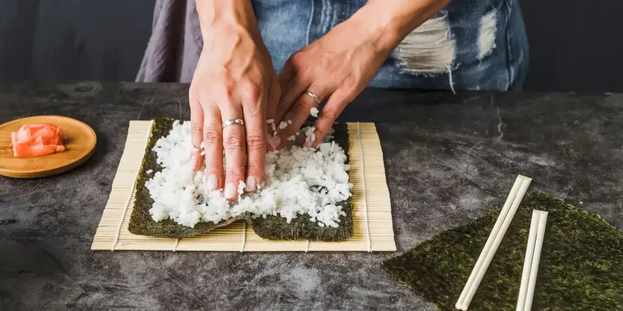 Dłonie rozprowadzają ryż na nori. Obok leżą pałeczki i marynowany imbir. To klucz do idealnego ryżu do sushi, który potrzebuje odrobiny octu.