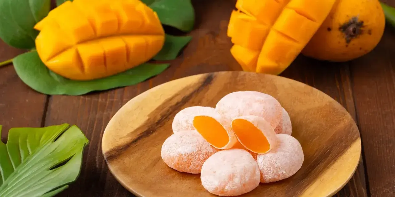 Słodkie mochi o smaku mango, ułożone na drewnianym talerzu, obok świeżych owoców i liści.